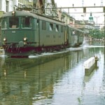 Die Re 4/4 I 10030 verlässt mit ihrem Personenzug den Bahnhof Rorschach Hafen. Offenbar haben sich die Bremsklötze und Radreifen des ersten Wagens ziemlich erhitzt. Die Wärme reicht jedenfalls um Wasser zu verdampfen. Die hoch liegende Rampe des Güterschuppens ist bevölkert mit Schaulustigen, die gut vor dem Wasser geschützt sind (von oben und unten). 01.08.1987 Foto: E. Meier